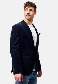 Marineblå cordfløyel blazer med skreddersydd passform, notched revers, to frontlommer og knappdetaljer på ermene. Båret over en hvit T-skjorte.