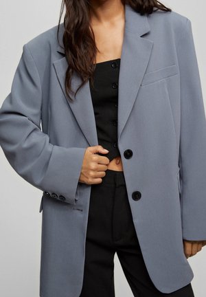 Femme portant un blazer gris oversize sur un crop top noir boutonné et un pantalon noir, tenant le blazer fermé d'une main.