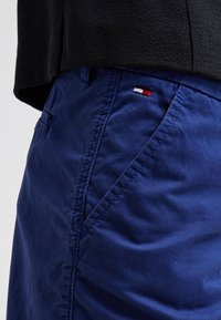 Detailní záběr na osobu v modrých kalhotách s šikmou přední kapsou a malým červeným, bílým a modrým logem značky, v kombinaci s černým strukturovaným topem.