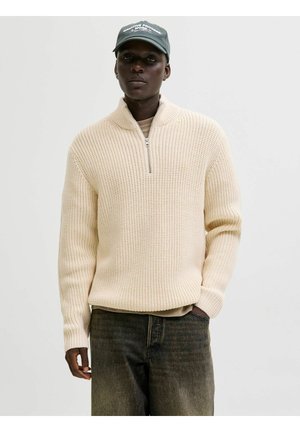 Nertinis užtrauktuku megztinis kreminės spalvos, su tekstūruotu raštu ir siaurais rankogaliais. Dėvimas virš šviesiai rudos palaidinės, kartu su tamsiai mėlynais džinsais ir žalia kepure.