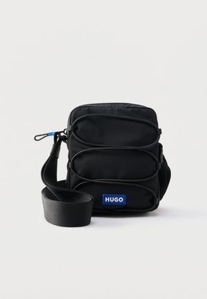 Compatta borsa a tracolla nera con tracolla regolabile, anelli elastici frontali e patch con logo "HUGO" blu su sfondo semplice.
