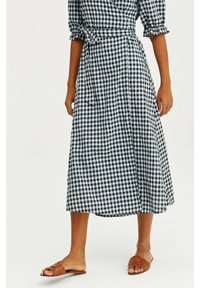 Femme portant une robe midi à carreaux noire et blanche avec des manches bouffantes et des sandales plates marron sur fond blanc.