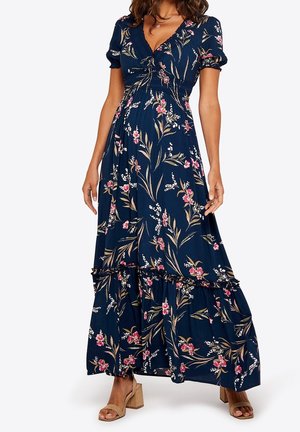 Kobieta w granatowej, kwiatowej sukience maxi z bufkami oraz beżowych sandałach na słupku, stojąca na tle jednolitego tła.