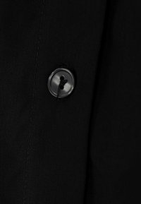 Schwarzer Stoff mit glatter Textur, ausgestattet mit einem schwarzen Kunststoffknopf. Der Knopf hat zwei Löcher und ist an dem Stoff angenäht.