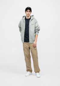 Sivi dukserica sa zipom i kenguruljim džepovima, crna majica ispod, kaki cargo hlače i bijele tenisice. Kapa upotpunjuje outfit.