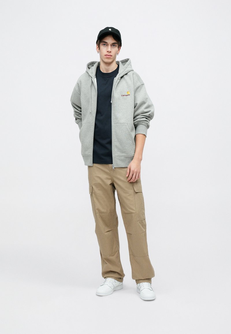 Felpa grigia con zip e tasche a marsupio, t-shirt nera sotto, pantaloni cargo color khaki e sneakers bianche. Il cappellino completa l'outfit.