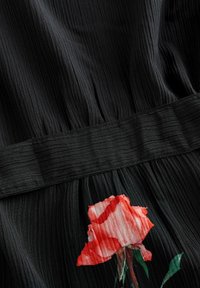 Zwarte geplooide stof met een textured oppervlakte; heeft een rood rozenpatroon aan de onderkant en een gladde zwarte tailleband erboven.