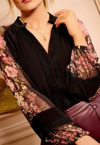 Blusa nera con maniche trasparenti a fiori e dettagli in pizzo. Presenta una chiusura con bottoni, scollatura rotonda e sovrapposizioni. Indossata con una borsa color porpora.