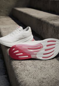 Białe sportowe buty z teksturowaną cholewką, różową podeszwą środkową i wzorzystą gumową podeszwą z różowymi i białymi akcentami.