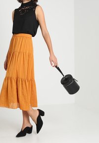 Nainen, jolla on yllään hihaton musta pitsitoppi, oranssi kerroksellinen midihame, mustat korkeat nilkkurit ja joka pitää mustasta sylinterimäisestä käsilaukustaan kiinni sen hihnasta.