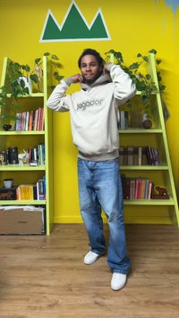 Κρεμ-χρώματος φούτερ με κουκούλα και λογότυπο, φορεμένο με μπλε τζιν και λευκά παπούτσια. Στο φόντο υπάρχουν πράσινα ράφια, βιβλία και κίτρινος τοίχος.