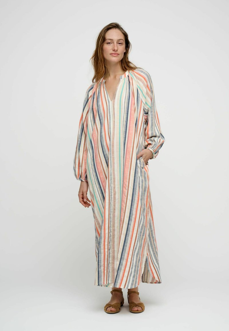 Femme debout portant une longue robe ample rayée aux tons pastels et des sandales, une main dans la poche, sur un fond clair uni.