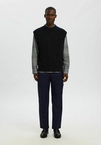 Tamsiai mėlynas megztinis be rankovių, dėvimas ant pilkos langinės ilgomis rankovėmis marškinio, derinamas su tamsiomis kelnėmis ir juodais bateliais. Neutrali fono.