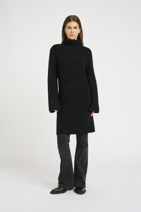 Abito nero a maglia con collo alto, maniche lunghe e taglio dritto. Abbinato a pantaloni neri a zampa e stivali. Dettagli minimi.