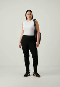 Vrouw in een witte mouwloze top, zwarte leggings, zwarte sneakers en met een zwarte schoudertas tegen een eenvoudige lichte achtergrond.