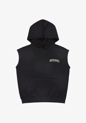 Czarna bezrękawnikowa bluza z kapturem z przednią kieszenią, biała koszulka pod spodem, jasnoniebieskie dżinsy i białe trampki. Kręcone włosy, stojąca poza.