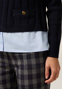 Navy-blauer, gerippter Strickpullover mit einer schwarzen Tasche und einem goldenen Knopf, über einem hellblauen Hemd und karierten Hosen mit braunen Akzenten getragen.