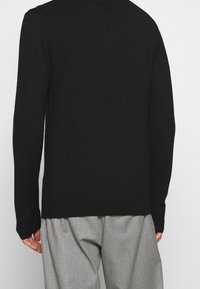 Schwarzer gestrickter Pullover mit V-Ausschnitt, langen Ärmeln sowie gerippten Bündchen und Saum; kombiniert mit grauen Nadelstreifen-Hosen.