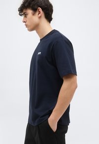 Νέος άνδρας με μπλε σκούρο T-shirt και μαύρο παντελόνι στέκεται προφίλ με το ένα χέρι στην τσέπη του, μπροστά σε απλό ανοιχτόχρωμο φόντο.