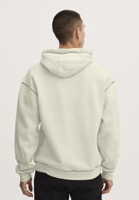 Sudadera gris clara hecha de tejido suave, con capucha ajustable, puños acanalados y un corte relajado. Detalle de costura visible a lo largo de las costuras.