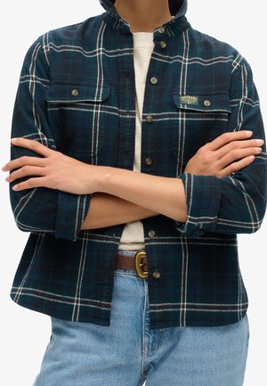 Personne portant une chemise à carreaux verts foncés et bleus avec poches poitrine, les bras croisés, un haut beige clair, un jean bleu et une ceinture marron.