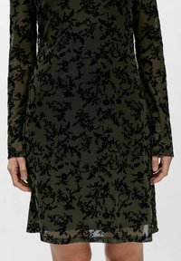 Moteris vilkinti alyvuogių žalios permatomos suknelės su ilgomis rankovėmis ir juodais aksominio gėlių raštais, matoma nuo vidurinės šlaunies iki torso.