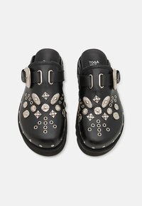 Sabots en cuir noir avec des accents décoratifs métalliques et des clous. Comprend deux boucles réglables et un design à bout rond.