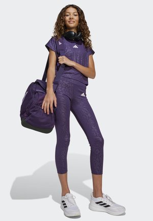 Jeune femme en tenue de sport violette et en baskets blanches, portant un sac de sport assorti et des écouteurs noirs autour du cou.