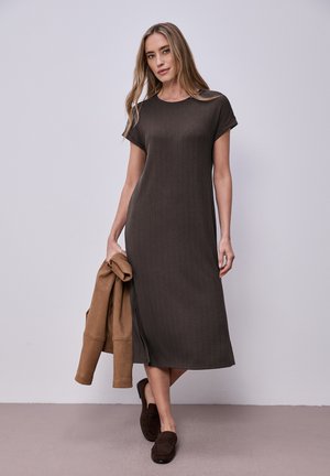 Kvinde står mod en ensfarvet væg iført en ribbet mørkebrun midi-kjole, holder en tanfarvet jakke og bærer mørkebrune loafers.