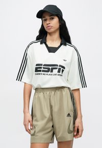 Bela ohlapna majica s črnimi poudarki in logotipom, v kombinaciji s bež kratkimi hlačami s stranskimi črtami in logotipom ter črno kapo.