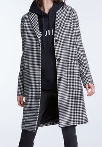 Mujer vistiendo un abrigo de patrón de pata de gallo en blanco y negro sobre una sudadera negra y pantalones negros, de pie contra un fondo liso.