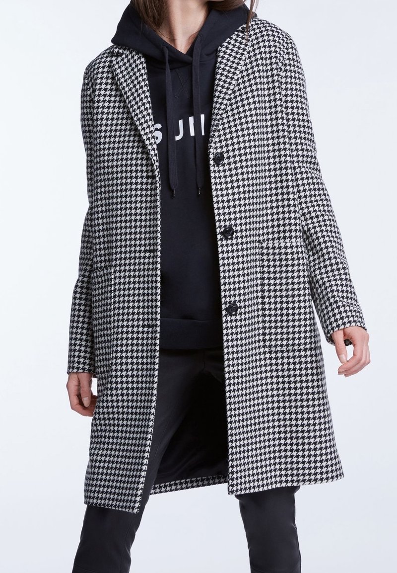 Mujer vistiendo un abrigo de patrón de pata de gallo en blanco y negro sobre una sudadera negra y pantalones negros, de pie contra un fondo liso.