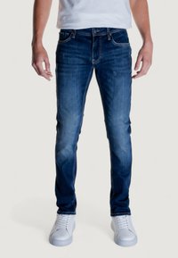 Jeans de mezclilla azul con un corte ajustado, que presentan un lavado desgastado, sutiles marcas de roce y un diseño clásico de cinco bolsillos. Combinados con zapatillas blancas.
