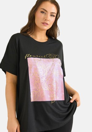 Frau mit langen braunen Haaren trägt ein schwarzes T-Shirt mit einem pinken Quadrat und floralem Gold-Design, hält den Saum des Shirts vor weißem Hintergrund.