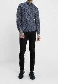 Camicia button-up blu navy con un piccolo motivo floreale bianco, maniche lunghe e colletto, abbinata a pantaloni neri slim-fit e scarpe nere.