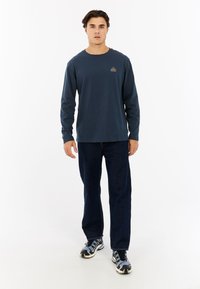 Marineblauw shirt met lange mouwen en een ronde hals, met een klein gouden logo op de borst, gecombineerd met donkere spijkerbroek en gemêleerde sneakers.