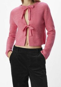 Cardigan en tricot rose au design court, avec une ouverture frontale nouée avec des nœuds, texture côtelée et manches longues. Associé à un pantalon en velours côtelé noir.