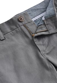 Pantalon gris avec fermeture éclair en laiton partiellement ouverte, bouton visible marqué « Authentic Next Dude » et doublure intérieure de ceinture rayée bleue.