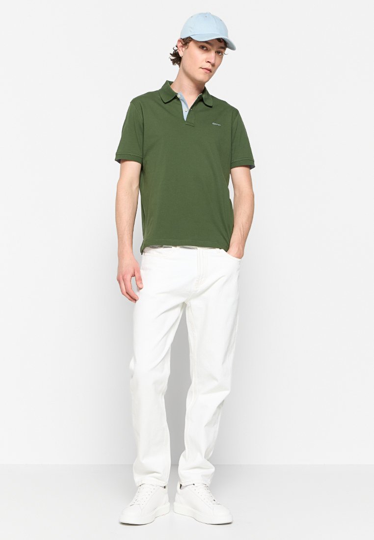 Polo vert avec un col bleu clair, assorti à un jean blanc et des baskets blanches. Le modèle porte une casquette bleu clair et adopte une posture décontractée.
