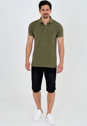 Polo de color verde oliva con mangas cortas y cuello, combinado con pantalones cortos de mezclilla negros y zapatillas blancas, posando sobre un fondo neutro.