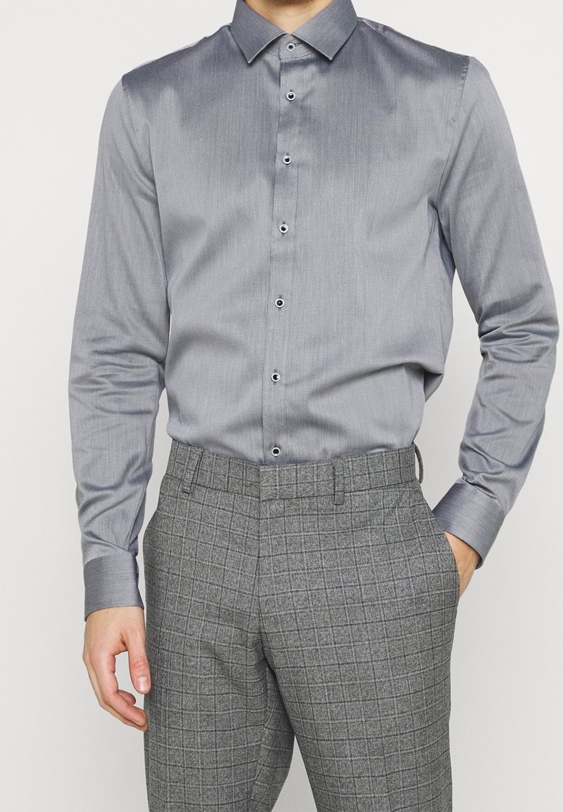 Homme portant une chemise grise ajustée avec des boutons noirs et un pantalon gris à carreaux, debout avec une main dans la poche.