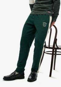 Dunkelgrüne Jogginghose mit einem weißen Seitenstreifen, versehen mit dem "SUPERDRY"-Logo in Weiß. Hergestellt aus weichem Material mit einer lockeren Passform und elastischem Bund.