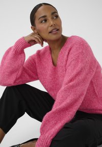 Suéter rosa esponjoso con cuello en V y mangas sueltas y texturizadas. El modelo está sentado con pantalones negros, mostrando la tela de punto suave.