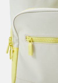 Närbild av en väska med en krämig yta och en gul dragkedja och detaljer, som visar en stilren, modern design.