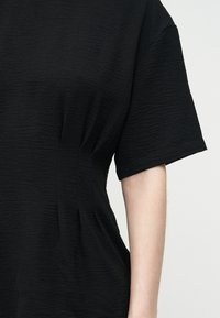 Μαύρο υφασμάτινο κοντομάνικο τοπ με λεπτομέρεια ραφής στη μέση στο πλάι, φορεμένο από ένα άτομο με ανοιχτόχρωμο μπράτσο που φαίνεται πάνω σε απλό φωτεινό φόντο.