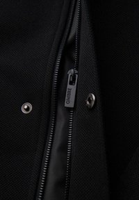 Zwarte, gestructureerde stof met een rits voorzien van een merkgetypt trekker. Zichtbare hardware omvat een drukknoop en metalen studs.