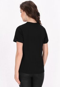 Sort bomulds T-shirt med opsmøgede korte ærmer, en klassisk pasform og en glat tekstur. Bæres med tætsiddende mørke bukser, set fra ryggen.