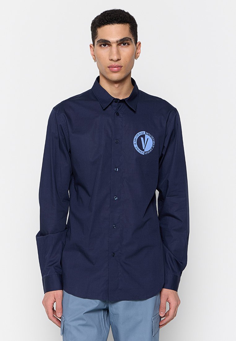 Chemise boutonnée en coton bleu marine avec un logo rond sur le côté gauche de la poitrine, col structuré et manches longues. Texture lisse, coupe standard.