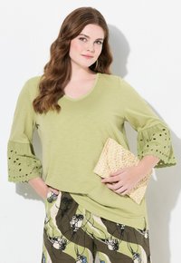 Maglietta verde chiaro a V con maniche a campana che presenta dettagli ad occhiello. Abbinata a dei pantaloni a fantasia e con una borsa a tracolla intrecciata.