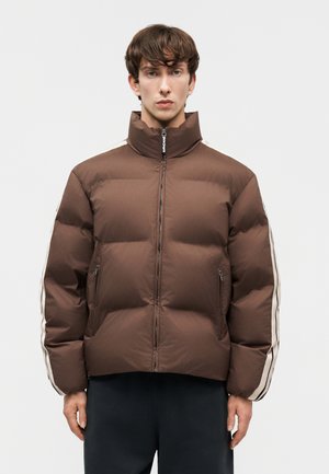 Młody mężczyzna zwrócony przodem, ubrany w brązową kurtkę puchową z białymi paskami na rękawach, z rękami luźno opuszczonymi wzdłuż boków.